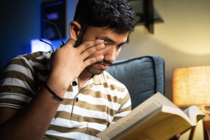 Close up shot young student with spectacles rubbing eyes due to eyes strain while reading book during late night - concept of feeling tired, early morning study and education